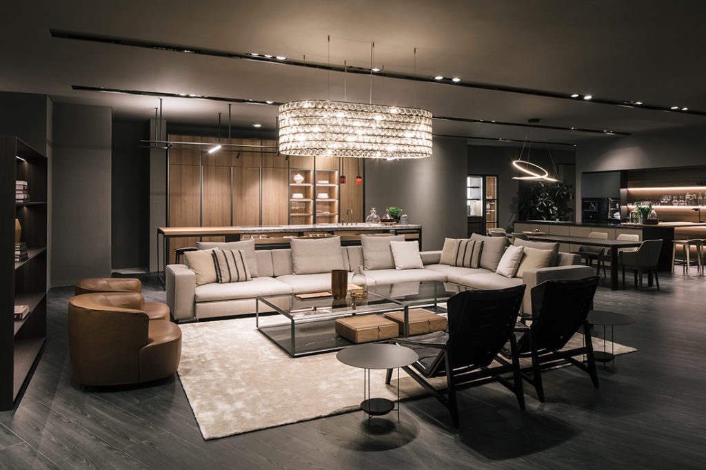 A sophisticated and dimly lit furniture showroom display showcasing a large modular sectional sofa, brown leather armchairs, glass-topped coffee tables, and an elegant crystal chandelier luxury furniture showrooms in Singapore.