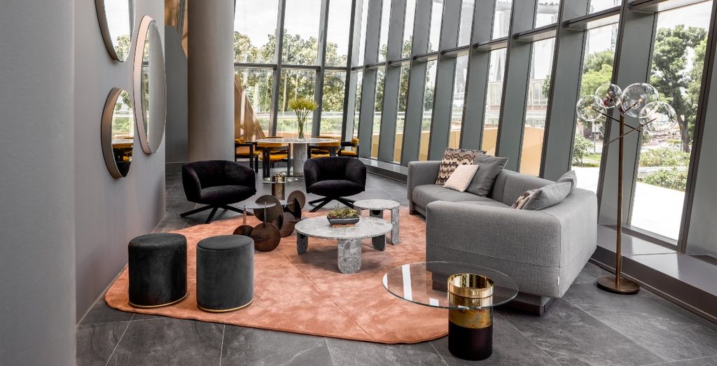 A stylish lounge area next to floor-to-ceiling windows with city views, featuring a grey sofa, black armchairs, round coffee tables, and a pink curved rug luxury furniture showrooms in Singapore.