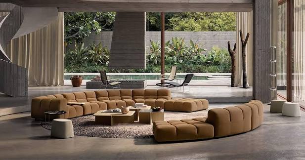 A modern, expansive open-plan living room with a large tan modular sectional sofa surrounding low wooden coffee tables, set against a backdrop of floor-to-ceiling glass windows overlooking a lush outdoor pool and tropical greenery luxury furniture showrooms in Singapore.