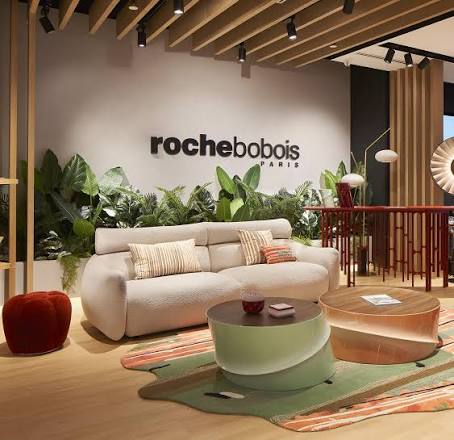 A modern furniture showroom featuring a light beige sofa with the "rochebobois" logo on the wall behind it, surrounded by indoor plants, colorful coffee tables, and wooden accents luxury furniture showrooms in Singapore.