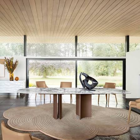 A bright, open-plan dining room with a large marble-topped table, modern chairs, a sculptural centerpiece, and large glass windows overlooking a green outdoor landscape under a wooden slated ceiling luxury furniture showrooms in Singapore.