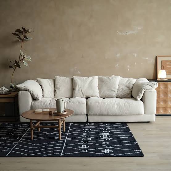 A contemporary living room featuring a large beige modular sofa with throw pillows, a small round wooden coffee table, a dark patterned rug, and a textured warm-toned wall luxury furniture showrooms in Singapore.
