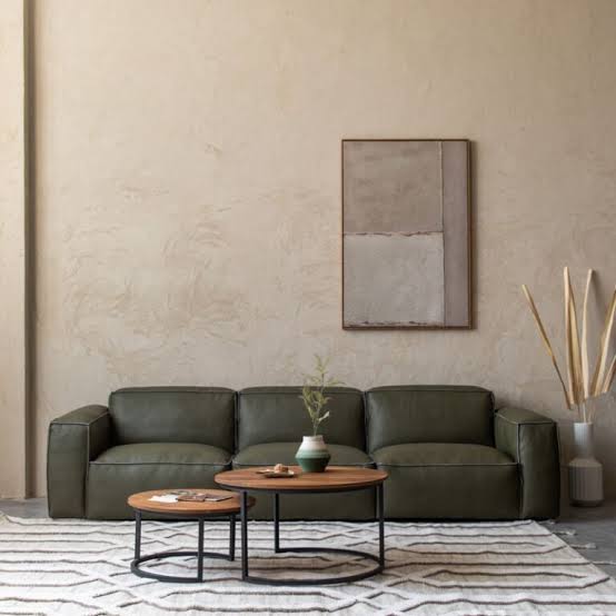 A minimalist living room setting with a dark green leather sofa, two nested round wooden coffee tables on a geometric rug, a textured beige wall, and a piece of abstract wall art luxury furniture showrooms in Singapore.