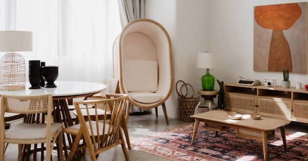 A brightly lit, modern dining and living room area featuring a round white dining table with wooden chairs, a unique enclosed pod chair, a low wooden coffee table on a patterned rug, and a media console against a white wall with abstract art luxury furniture showrooms in Singapore.