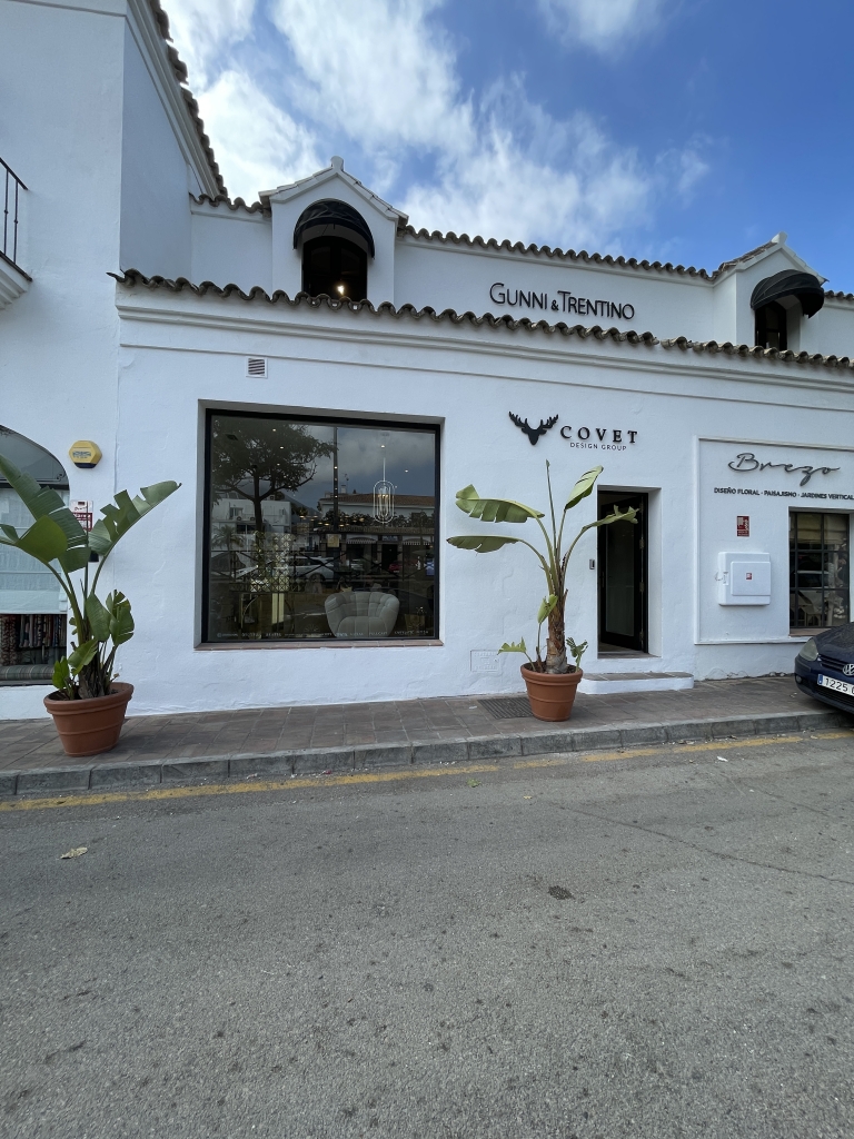 Marbella- Exterior view of a white building housing the Covet Design Group showroom. The facade features two large windows, one with visible modern furniture and a stylish chandelier inside. Two large potted plants with broad leaves flank the entrance. Above, the signs read "Gunni & Trentino" and "Covet Design Group". The street and sidewalk are visible in the foreground under a partly cloudy sky.
