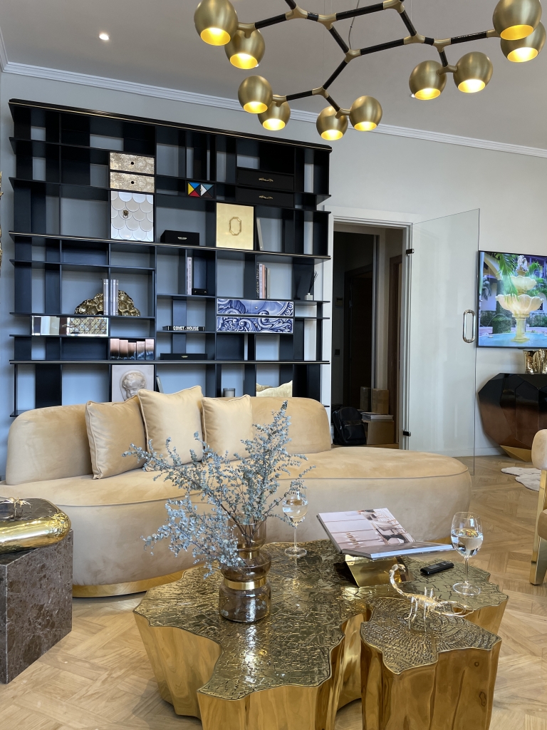 Marbella- Sophisticated living room interior featuring a curved beige velvet sofa with matching cushions, a unique gold sculptural coffee table with an organic, textured design, and decorative elements including a glass vase with dried flowers, magazines, and gold figurines. Behind the sofa is a large black shelving unit with artistic drawers and books. A modern chandelier with gold spheres hangs from the ceiling, and a wall-mounted TV is visible in an adjacent room through a glass door.
