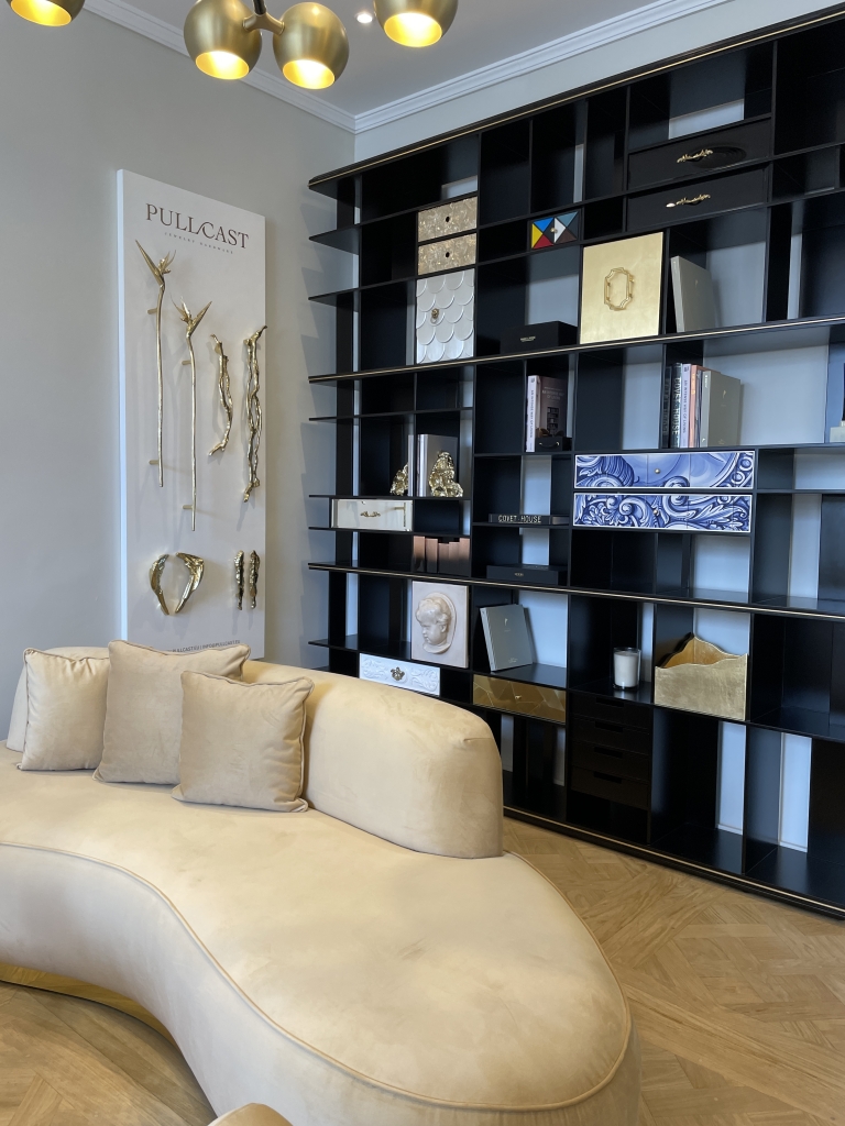 Marbella- Stylish and modern interior space featuring a sculptural beige curved sofa with matching cushions, placed in front of a large black open shelving unit filled with artistic decor, books, and drawer elements. A wall display by PullCast showcases luxurious gold hardware pieces shaped like branches and organic forms, under a contemporary gold light fixture.