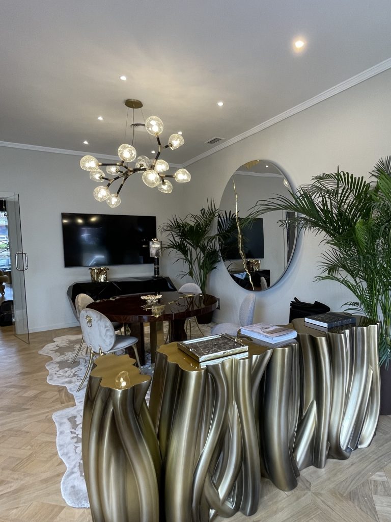 Marbella- Luxurious modern interior featuring a sculptural gold-toned console table with a fluid, organic design, paired with a round dining table and velvet chairs. A statement chandelier with multiple glass globes hangs from the ceiling. Large indoor plants, a circular wall mirror, and a flat-screen TV enhance the elegant ambiance.