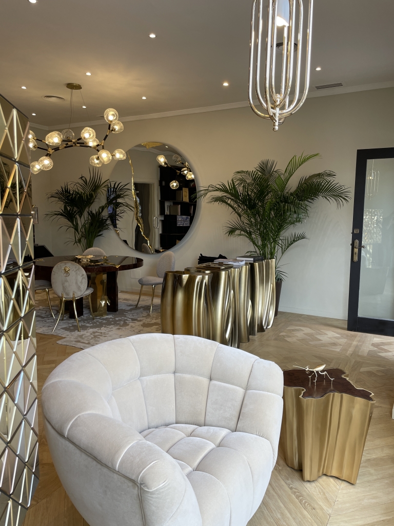 Marbella- Luxurious interior with a plush, curved cream armchair in the foreground, next to a gold sculptural side table. In the background, a dining area features velvet chairs with gold details, a round wooden table, and a dramatic chandelier with globe lights. A mirrored wall, tall indoor plants, and fluid gold-toned console tables add to the elegant and contemporary ambiance.