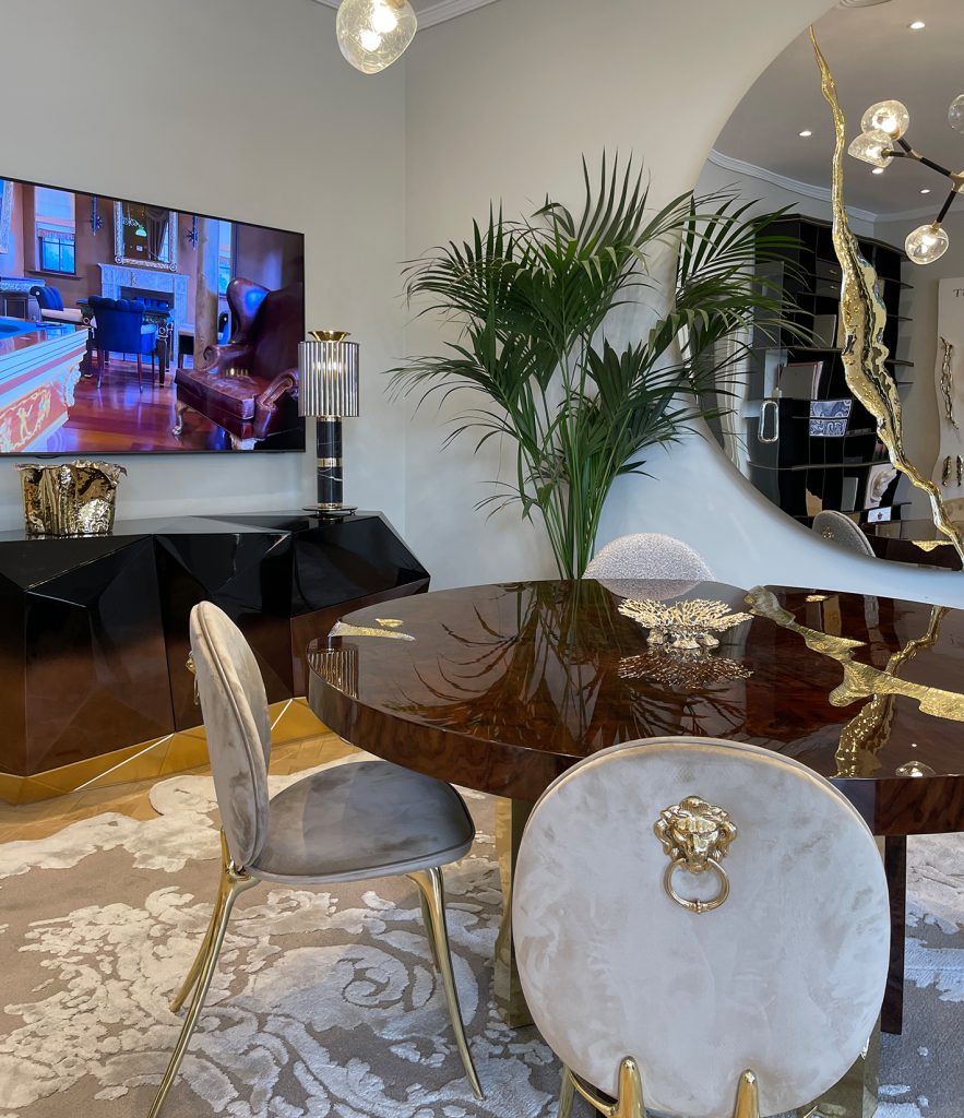 Marbella- Elegant dining area featuring a glossy round wooden table with gold inlays, surrounded by velvet chairs with gold lion head details on the backrests. A large decorative mirror, indoor plant, and flat-screen TV enhance the luxurious atmosphere, along with a sculptural black sideboard and gold-accented decor elements.
