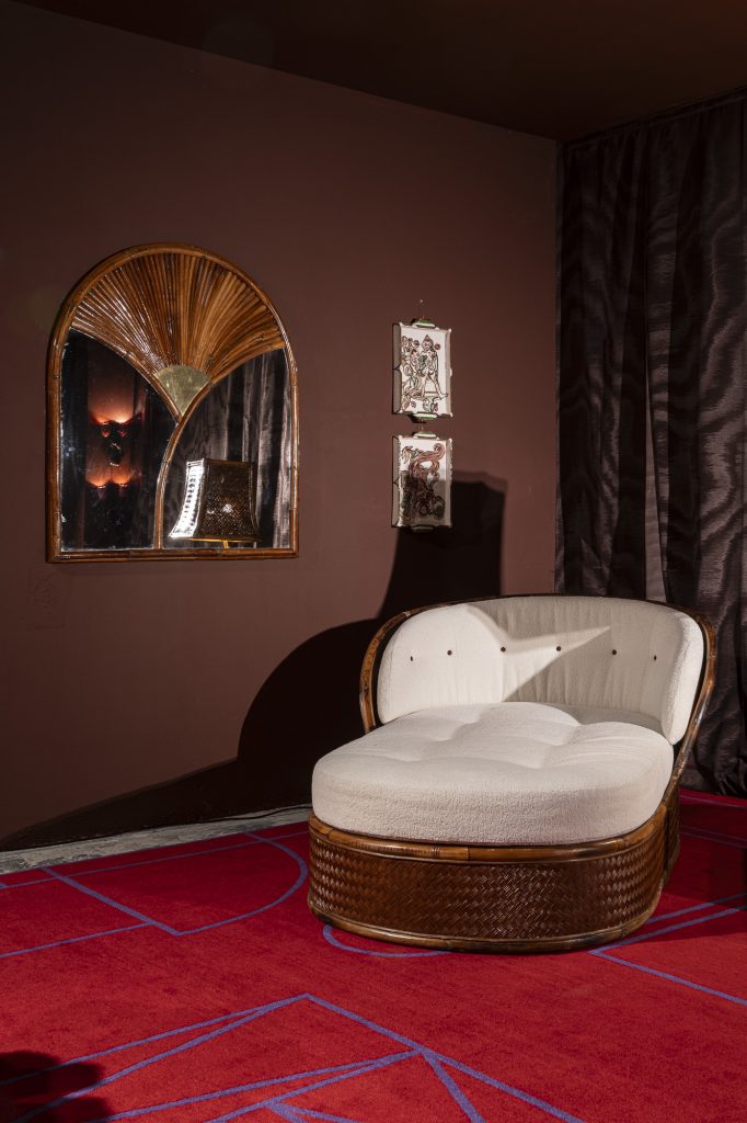 NOMAD St. Moritz 2026, A vintage bamboo chaise longue with white upholstery set against a chocolate brown wall with a sunburst-patterned rattan mirror and a geometric red rug.