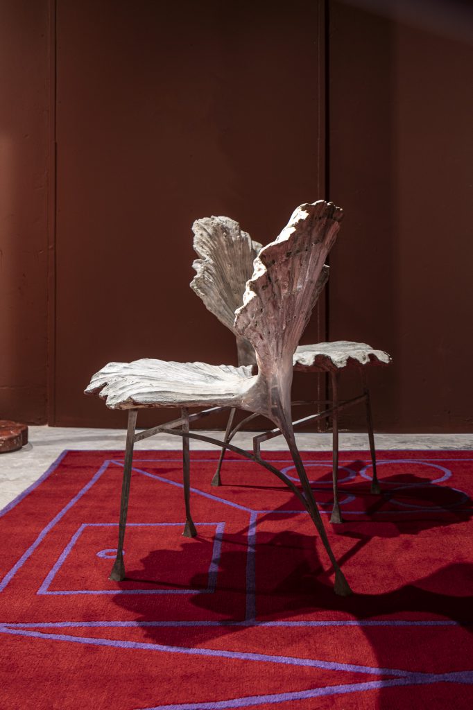 NOMAD St. Moritz 2026, Two organic-shaped cast metal chairs resembling large gingko leaves or shells positioned on a vibrant red carpet with purple geometric outlines.
