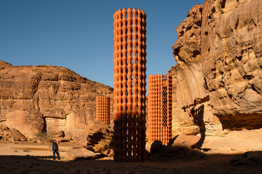 architecture events 2026, Tall pillars made of stacked terracotta clay pots standing in a vast desert canyon landscape with rock formations under a clear blue sky.