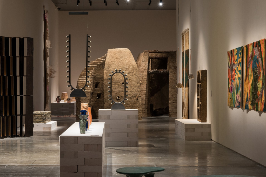 architecture events 2026, Spacious art gallery featuring earthen adobe-style sculptures, modern lighting fixtures, and colorful abstract textile art on white walls.
