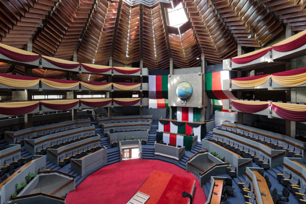 architecture events 2026, Interior of a large Brutalist-style parliamentary assembly hall with a geometric wood-paneled ceiling, red carpets, and tiered seating.