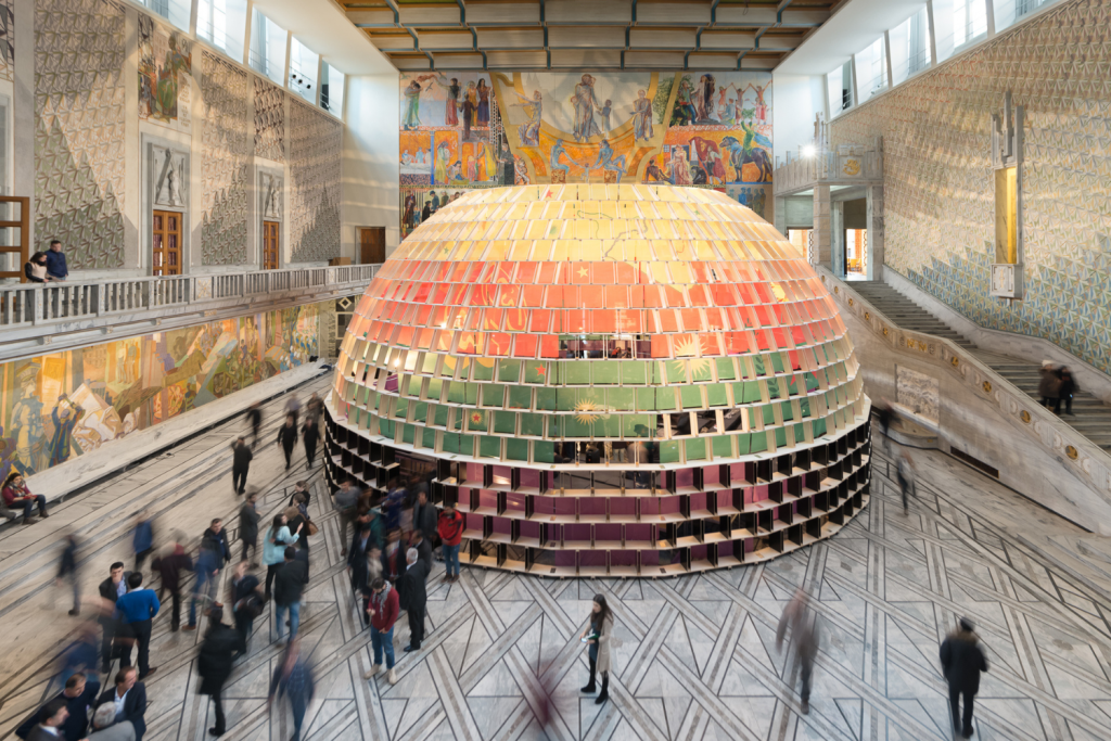 architecture events 2026, A large, multi-colored geodesic dome structure installed in the center of a grand marble hall with intricate wall murals and people walking by.