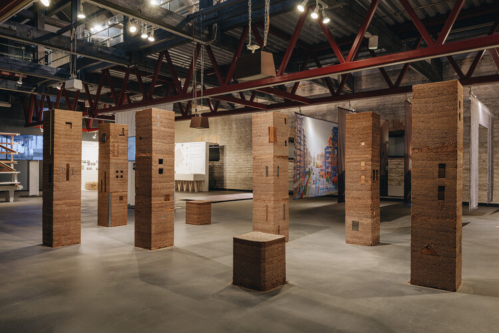 architecture events 2026, Exhibition space featuring tall rectangular columns made of rammed earth and natural materials under industrial ceiling trusses.
