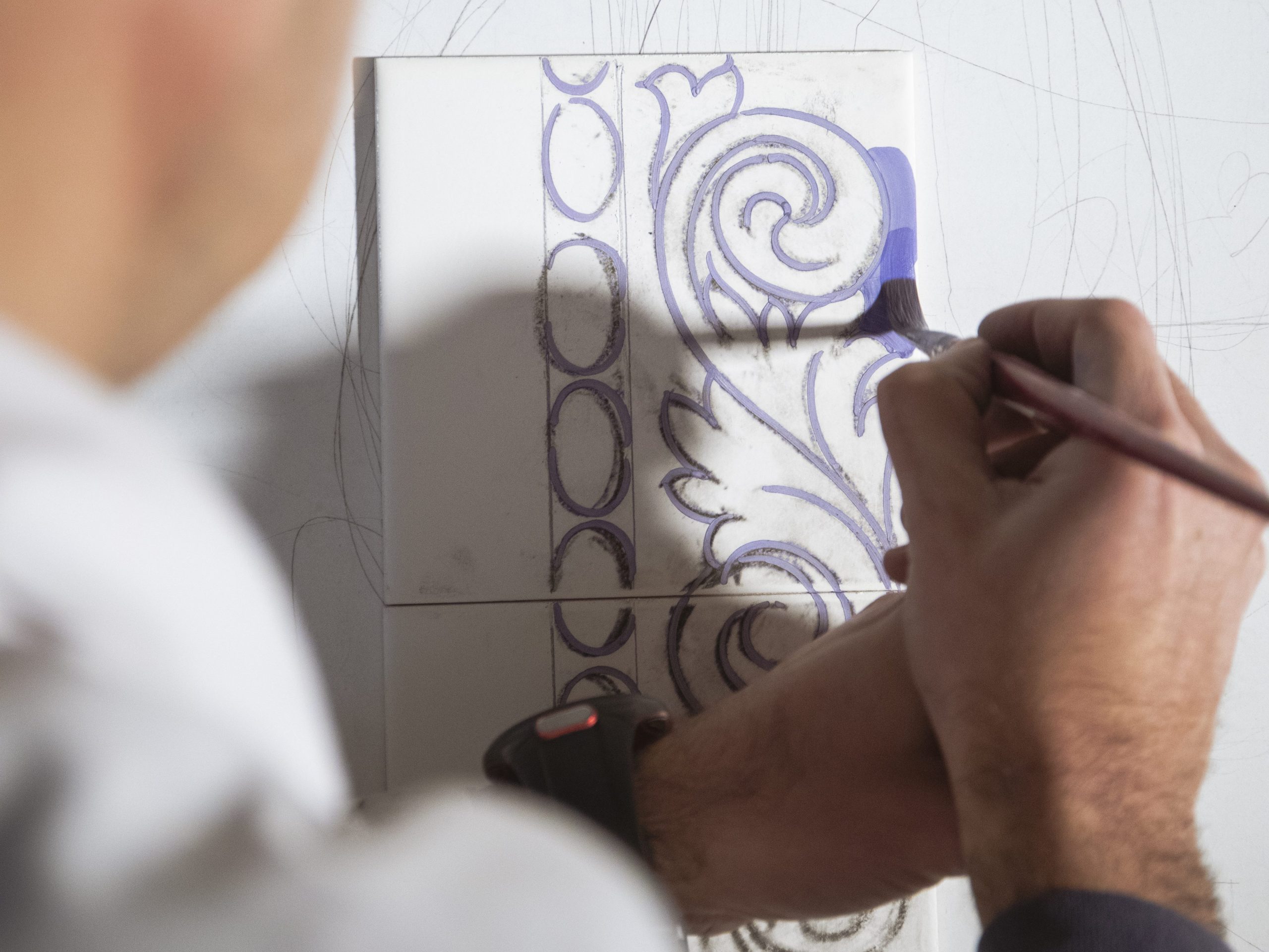Close-up of an artist's hands using a brush to paint a purple ornamental design on a white ceramic tile, Quarry Tiles Center Table