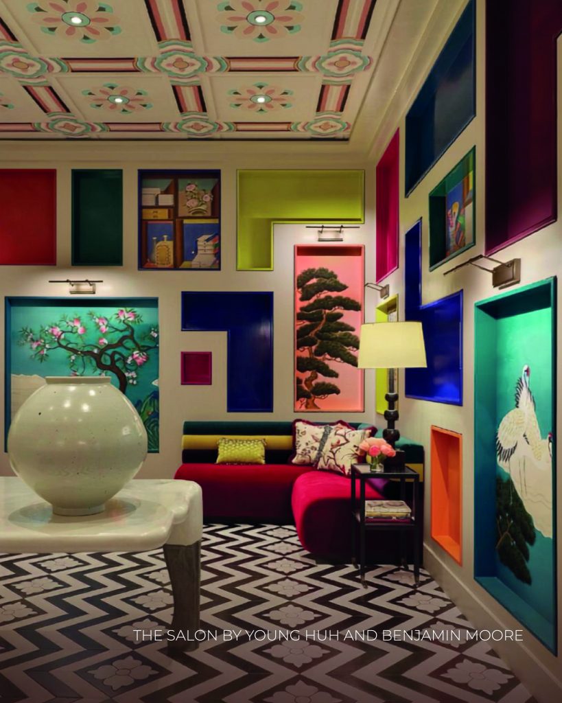 Sala de estar moderna e eclética com forte uso de cores primárias e formas geométricas. As paredes brancas apresentam vários nichos embutidos em formatos retangulares e em "L", pintados em cores vibrantes como azul-escuro, vermelho, amarelo, verde e laranja; alguns destes nichos contêm obras de arte de estilo oriental, como painéis com árvores e garças. O teto em caixotão exibe pinturas circulares florais e iluminação embutida. Um sofá de canto modular em veludo vermelho-vivo está encostado à parede, e o chão é revestido por um tapete com um marcante padrão ziguezague em preto e branco com pequenos detalhes florais. Em primeiro plano, uma mesa branca brilhante exibe um grande vaso esférico de cerâmica clara. Na base da imagem, lê-se o texto "THE SALON BY YOUNG HUH AND BENJAMIN MOORE". WOW!house