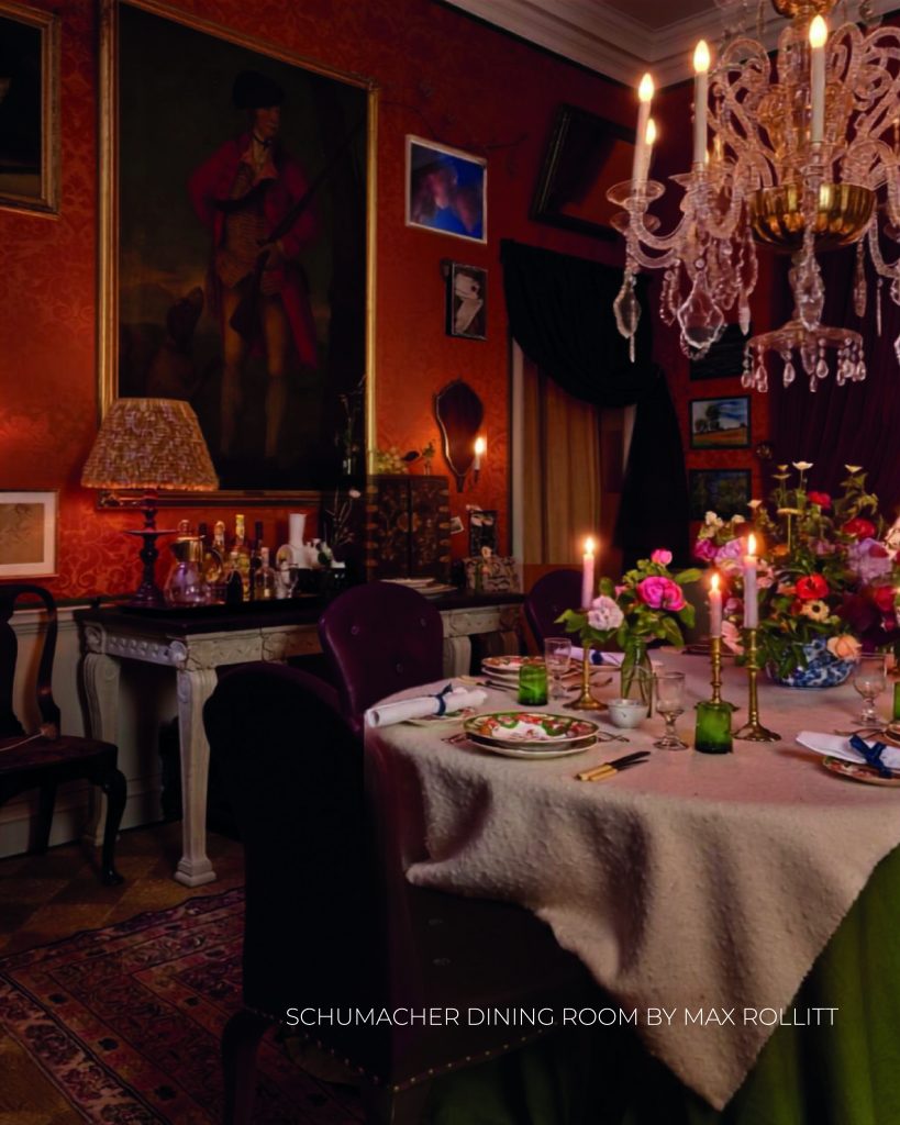 Sala de jantar formal e clássica com iluminação quente à luz de velas. Uma grande mesa redonda coberta com uma toalha texturizada de tom claro está posta com pratos decorados, copos verdes e vários castiçais com velas acesas. Um sumptuoso lustre de cristal pende sobre a mesa, refletindo a luz. As paredes ao fundo são de um tom vermelho-alaranjado quente com damasco subtil, decoradas com quadros emoldurados, incluindo um grande retrato a óleo de uma figura histórica com trajes vermelhos. Um aparador de madeira clara com garrafas de bebidas e um candeeiro de mesa com abajur plissado completam o cenário clássico. Na base da imagem, lê-se o texto "SCHUMACHER DINING ROOM BY MAX ROLLITT". WOW!house