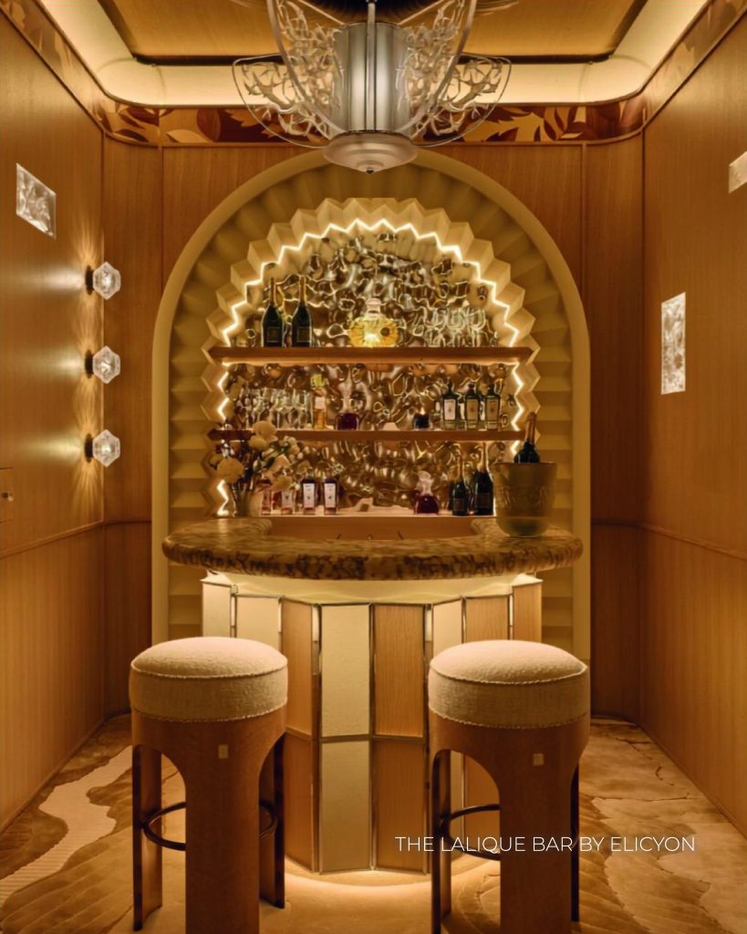 Um bar compacto e luxuoso com design geométrico e tons dourados e amadeirados. O balcão do bar é curvo, feito de mármore veteado escuro, acompanhado por dois bancos altos com assentos redondos e estofados a branco e pernas de madeira arqueadas. Atrás do balcão, um nicho em arco iluminado exibe prateleiras com garrafas sobre um fundo texturizado e espelhado que brilha intensamente com fitas de LED ocultas. As paredes laterais são revestidas com painéis de madeira clara e decoradas com candeeiros de parede geométricos em cristal esculpido. Na base da imagem, lê-se o texto "THE LALIQUE BAR BY ELICYON". WOW!house