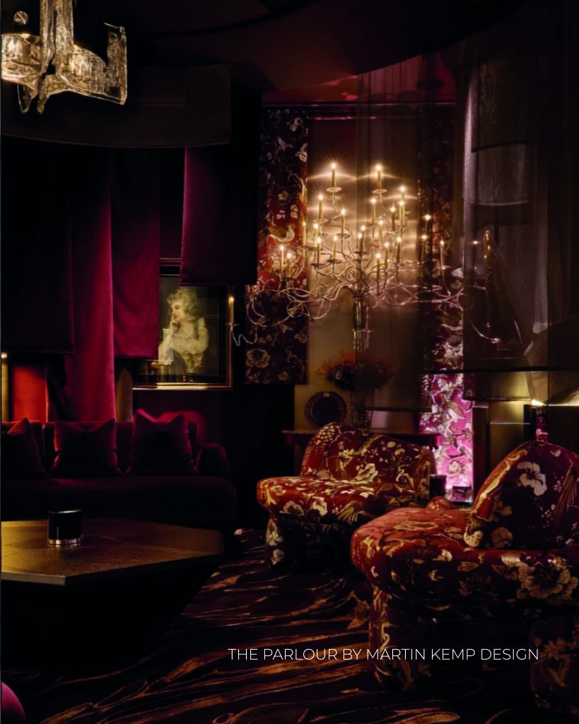 Sala de estar com ambiente intimista, dramático e iluminação cenográfica. O espaço destaca-se por um grande lustre ramificado com múltiplas lâmpadas acesas que criam um brilho intenso. Duas poltronas robustas em primeiro plano estão estofadas com um tecido de padrão floral maximalista em tons de vermelho, dourado e preto. Ao fundo, um sofá escuro está posicionado sob cortinas de veludo vermelho-escuro pesadas e uma pintura clássica iluminada na parede. O chão exibe um tapete com padrão fluido e escuro, refletindo a iluminação dourada e quente do espaço. Na base da imagem, lê-se o texto "THE PARLOUR BY MARTIN KEMP DESIGN". WOW!house