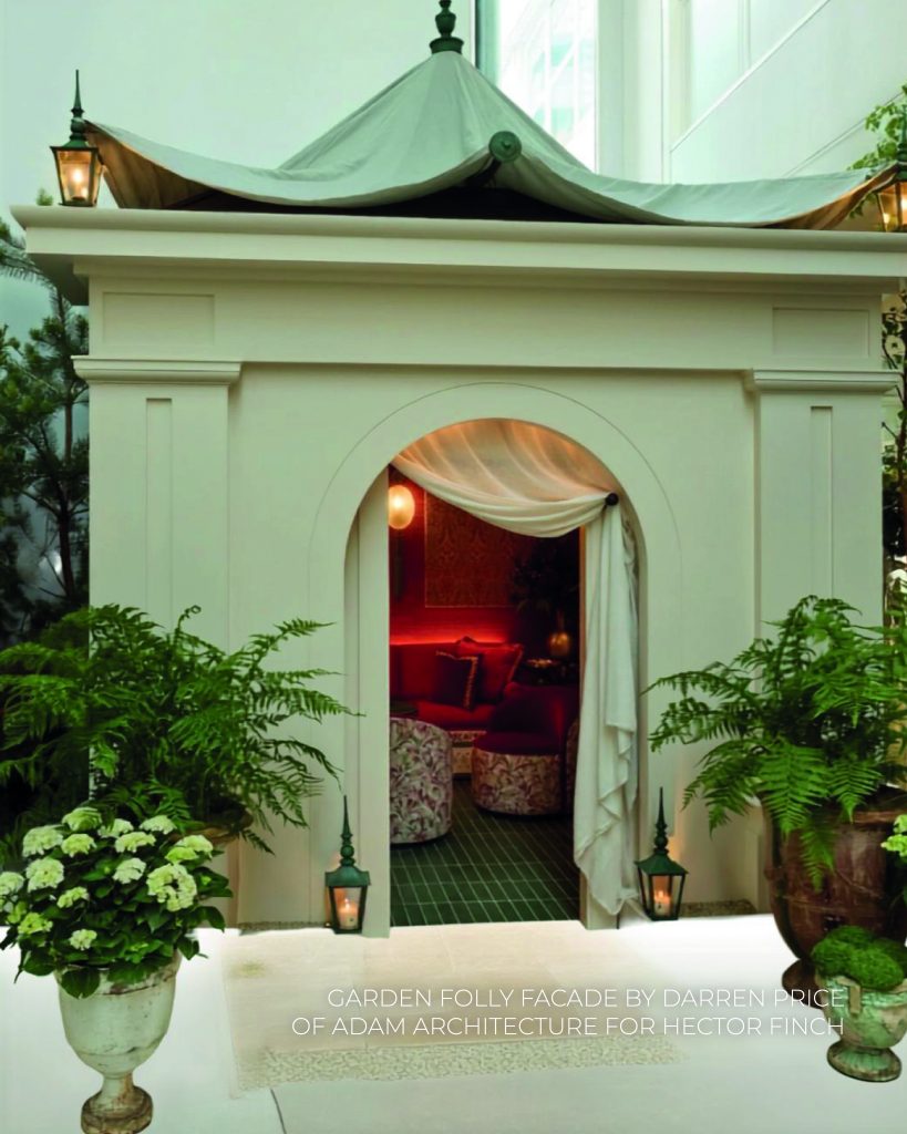 Vista detalhada do interior da "Garden Folly", uma sala luxuosa decorada inteiramente em tons intensos de vermelho e dourado. Um sofá de veludo vermelho vibrante preenche o fundo, decorado com várias almofadas texturizadas de veludo e cetim vermelho com franjas douradas. À frente do sofá, encontram-se duas poltronas curvas e baixas com estofo interior vermelho e exterior com um padrão abstrato em tons claros e avermelhados. As paredes são revestidas de tecido vermelho texturizado com grandes painéis verticais em brocado dourado e candeeiros de parede ovais com borlas compridas. No centro, uma mesa de apoio redonda e verde com franjas serve de apoio. Na base da imagem, lê-se o texto "GARDEN FOLLY ROOM BY STUDIO ENASS". WOW!house