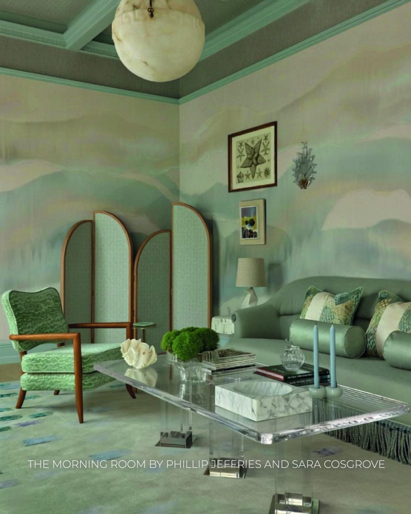 Sala de estar com uma atmosfera serena e tons suaves de verde e pastel. As paredes exibem uma pintura mural artística contínua que evoca montanhas enevoadas em tons suaves. No centro, destaca-se uma mesa de centro retangular em acrílico transparente com objetos decorativos, como velas azuis e uma concha branca. Um sofá cinzento-esverdeado com almofadas estampadas e rolos decorativos está encostado à parede direita. No canto, um biombo de três painéis arqueados com padrão verde claro complementa o espaço ao lado de uma poltrona estofada em tecido aveludado verde com braços de madeira. Um grande candeeiro esférico de pedra translúcida pende do teto em caixotão cinzento-azulado. Na base da imagem, lê-se o texto "THE MORNING ROOM BY PHILLIP JEFFERIES AND SARA COSGROVE". WOW!house