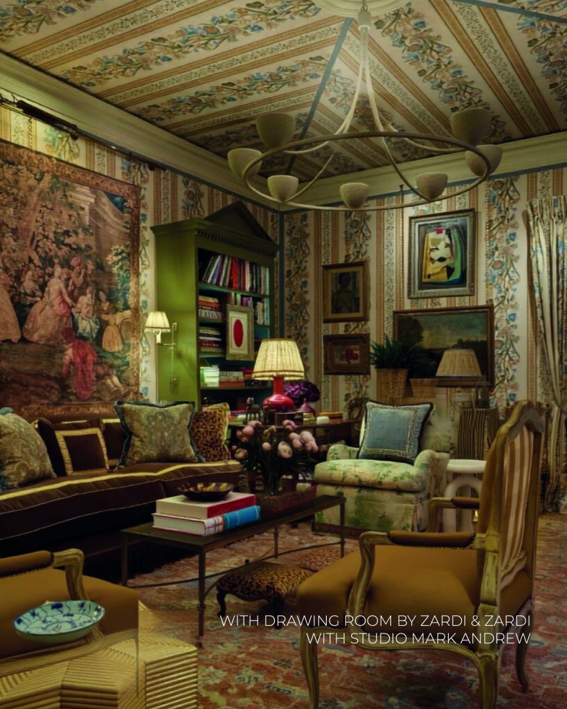 Sala de estar com decoração clássica, maximalista e opulenta. O teto é ornamentado com painéis pintados com motivos florais e exibe um grande lustre de metal em formato de anel. As paredes estão totalmente revestidas com papel de parede às riscas verticais decoradas com padrões botânicos. Do lado esquerdo, uma grande tapeçaria clássica adorna a parede acima de um sofá de veludo castanho-escuro repleto de almofadas decorativas. Uma estante de livros verde-oliva com topo em frontão destaca-se ao fundo. No centro, uma mesa de centro escura exibe livros empilhados e arranjos florais, rodeada por poltronas com diferentes estofos estampados e clássicos. Na base da imagem, lê-se o texto "WITH DRAWING ROOM BY ZARDI & ZARDI WITH STUDIO MARK ANDREW". WOW!house