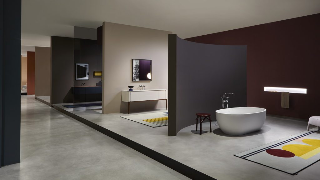 A sophisticated, architectural showroom for luxury bathroom design. The space is divided into a maze of intersecting walls in varying shades of dark beige and plum-red. Integrated into these structures are high-end display pieces: a sleek, curved bathtub with floor-mounted fixtures, a minimalist white vanity unit with a unique patterned mirror, and a linear wall light. A colorful, abstract-patterned rug with geometric shapes adds a burst of color. luxury showrooms