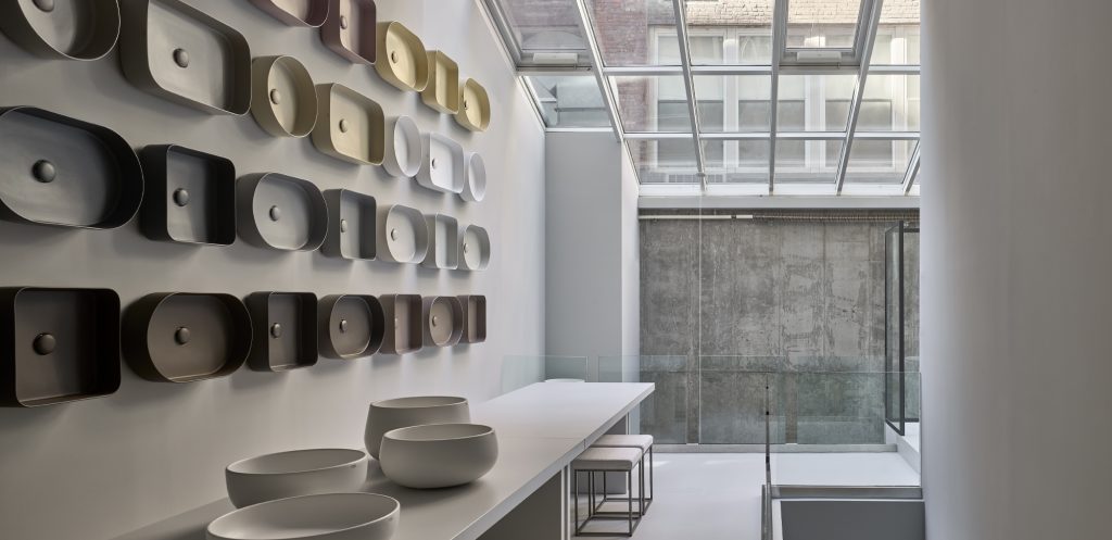 A sophisticated showroom for high-end bath fixtures. The space is flooded with natural light from a large, open glass skylight, exposing a raw, textured concrete wall. On a long, built-in display shelf, dozens of different colored and shaped basins are meticulously mounted. A white countertop features two sleek, rounded stone basins. The overall aesthetic is minimalist, with polished concrete floors and glass railings. luxury showrooms