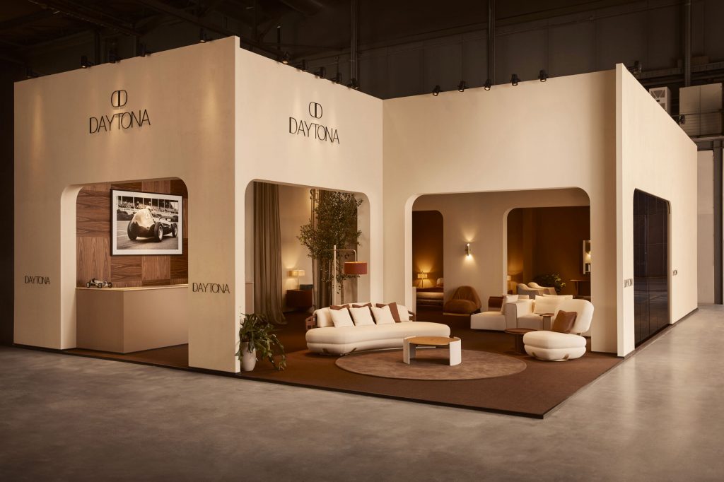 A brightly lit luxury brand stand named "Daytona" at a trade show. The stand has a clean, textured plaster finish and features multiple curved arches. One archway frames a reception area with a black and white framed photo of a vintage race car and a small model car on the counter. The other archway displays a modern furniture collection, including curved off-white sofas and a coffee table, surrounding a large indoor plant in a terracotta pot. luxury showrooms
