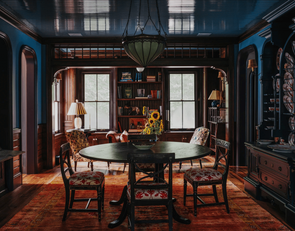 a cozy, traditional study with floor-to-ceiling dark wood shelving, a round central table on a patterned rug, blue high-gloss ceilings, and a massive glass chandelier. ELLE Decor A-List