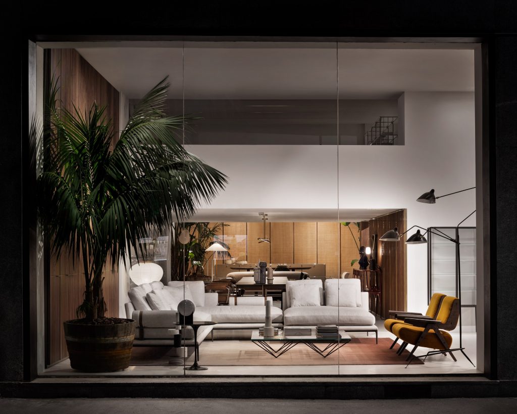 An exterior view through a large glass window shows a contemporary interior furniture showroom. A large potted palm stands to the left of an L-shaped light grey sofa, two yellow armchairs, and designer lamps, luxury showrooms.