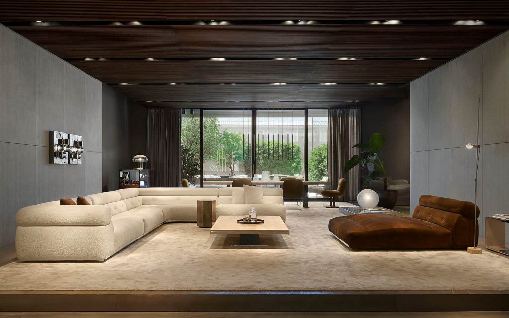 A photograph of a large interior space with a distinctive linear tube lighting grid on a dark ceiling. Modular sofas in teal, orange, and lime green are arranged in small vignettes across the space, bordered by high-standing pale wooden panel dividers, luxury showrooms.