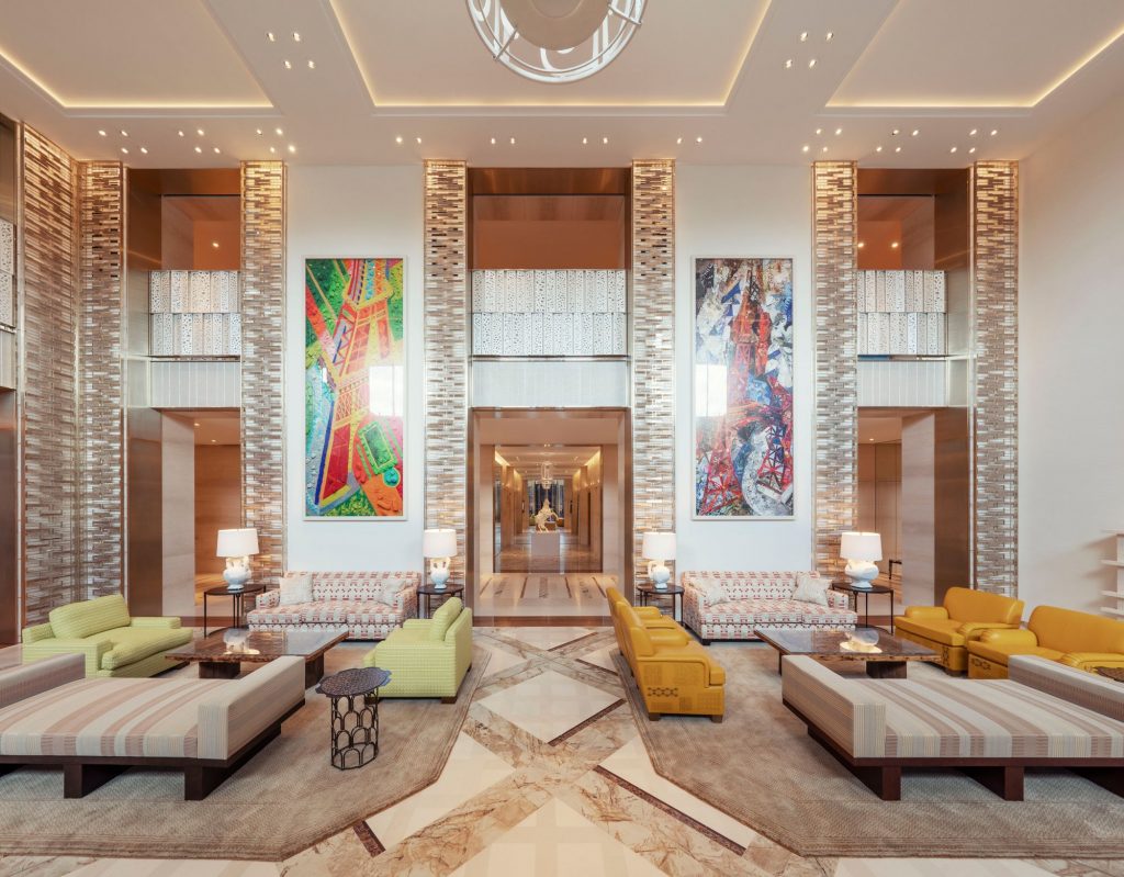 a grand, symmetric luxury hotel lobby with patterned geometric flooring, double-height columns, large-scale modern art, and matching low-slung modern benches and colorful sofas. ELLE Decor A-List