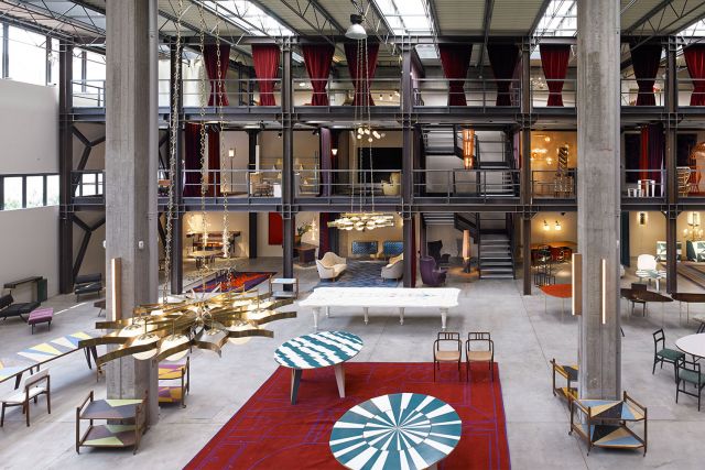 An elevated view looking down into a sprawling multi-level industrial design showroom. Steel framing with red velvet curtains defines the display areas, filled with diverse designer furniture pieces like multi-colored tables, various chairs, and unique seating arrangements, luxury showrooms.
