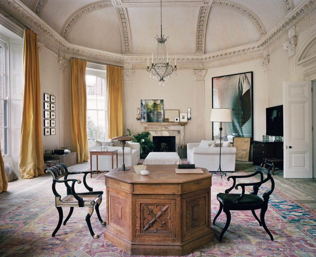 A high-ceilinged room with yellow curtains, a large central table, and artistic decor. ELLE Decor A-List