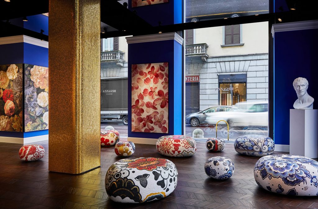 An immersive retail showroom for decorative glass and tile mosaics. A grand, square pillar covered in sparkling gold mosaic tiles dominates the space. Various round and oval poufs, meticulously decorated with intricate floral and geometric mosaic patterns (some blue and white, some red and white), are artfully arranged on a dark chevron-patterned wood floor. Large-scale mosaic art panels hang on deep blue walls, including one with pink blossoms. luxury showrooms