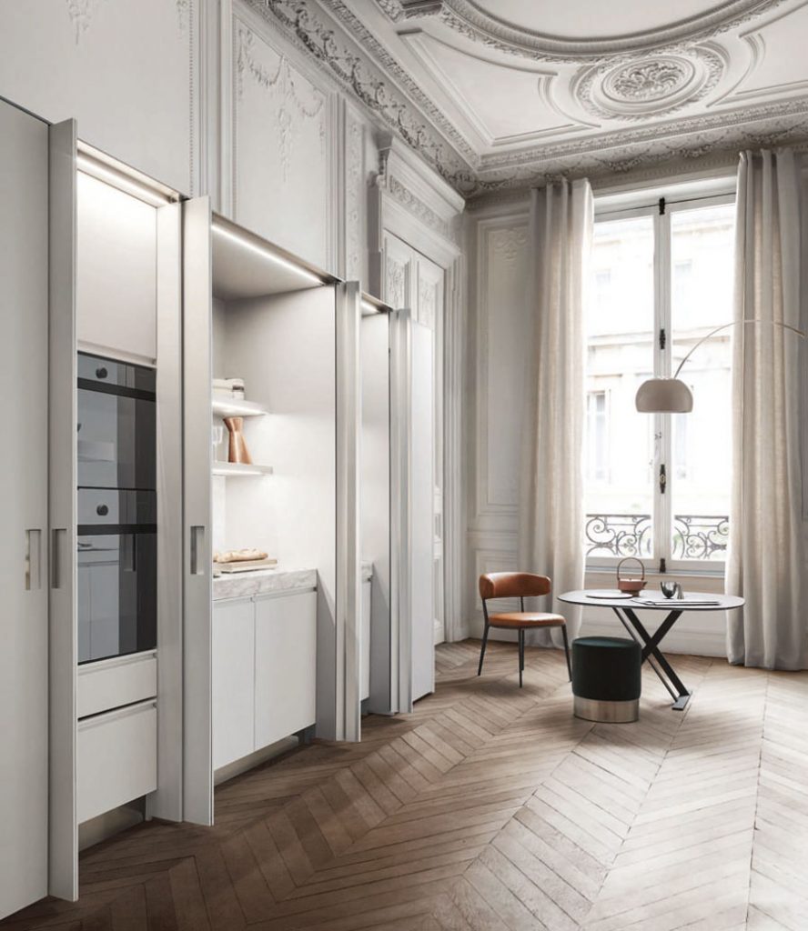 A perspective shot of a modern, open-plan kitchen and dining area set within a luxurious apartment featuring ornate ceiling stucco. Integrated white cabinetry with built-in appliances and lit shelving is visible, next to a black table with leather and velvet seating, luxury showrooms.