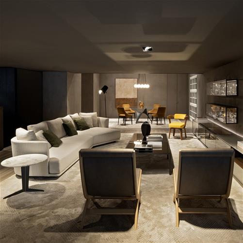 A large, modern, open-plan living and dining area in a luxury showroom. In the foreground, a curved, modular white sofa with olive green throw pillows is paired with a round marble-top side table. The space is illuminated by warm, recessed lighting and a striking modern chandelier over the dining table with mustard yellow chairs in the background. A large textured area rug covers the concrete floor. Built-in display cases with intricate glass pieces line the far wall. luxury showrooms