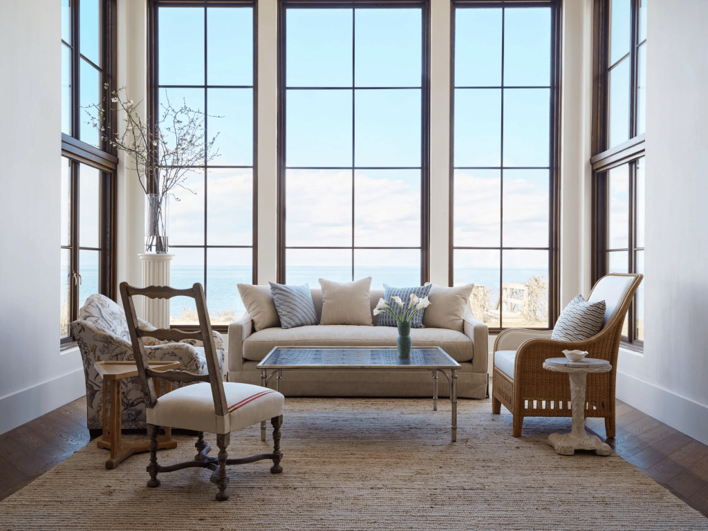 a bright sunroom overlooks the ocean through tall grid windows, featuring various natural-fiber and patterned armchairs centered around a glass and silver coffee table on a jute rug. ELLE Decor A-List