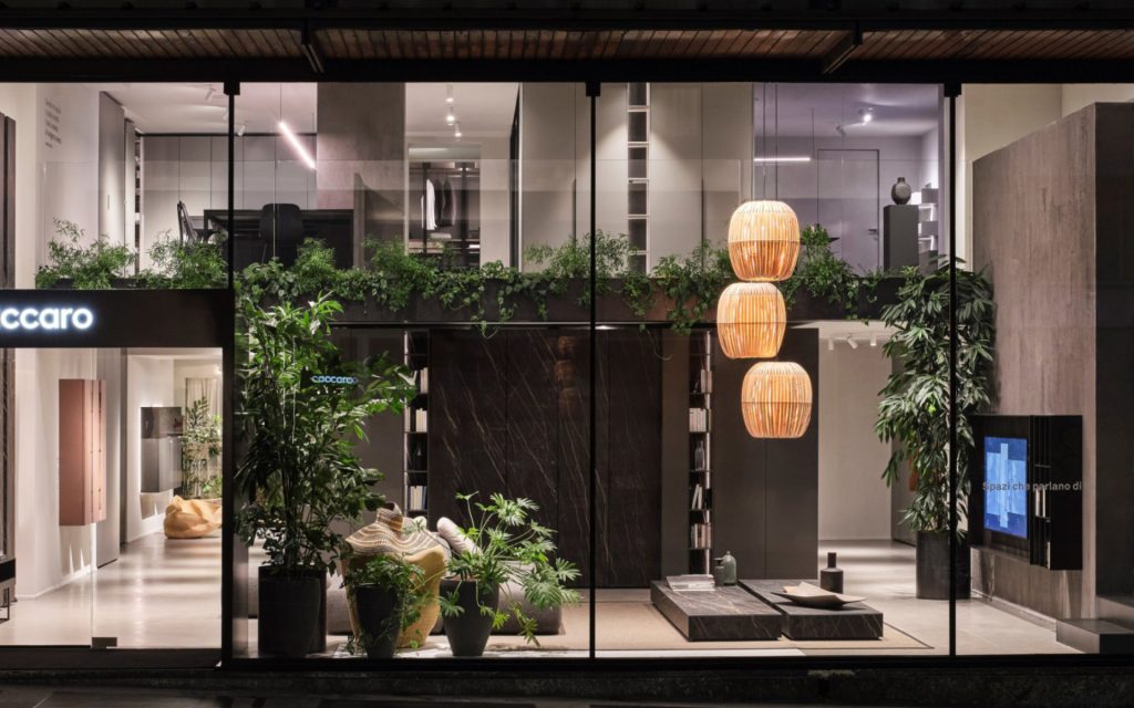 A stunning, modern, multi-level furniture showroom facade at night, viewed through a large glass window from the street. The exterior features built-in planter boxes with cascading greenery. Inside, a complex, architecturally intricate design is revealed. Two large, golden bamboo pendant lamps hang, casting warm light on a central dark marble-block coffee table and a selection of potted plants. The upper level has glass railings and a mix of display pieces. luxury showrooms