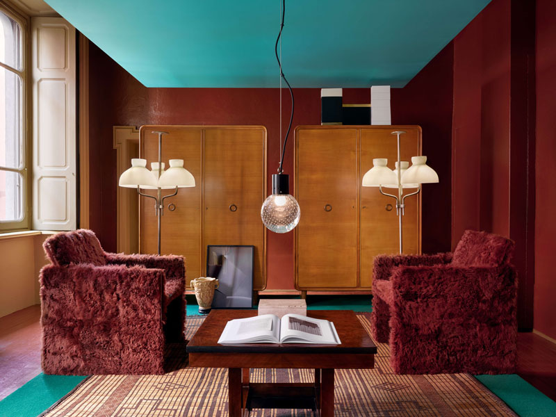 An eclectic and richly colored living room with deep terracotta walls and a vibrant turquoise ceiling. Two plush, shaggy maroon armchairs face a vintage wooden coffee table with an open book. A patterned woven rug is on the floor, and in the background, two matching wooden cabinets stand next to unique multi-globe floor lamps. An artistic pendant light hangs from the ceiling. A framed artwork and a decorative vase rest on the coffee table. luxury showrooms