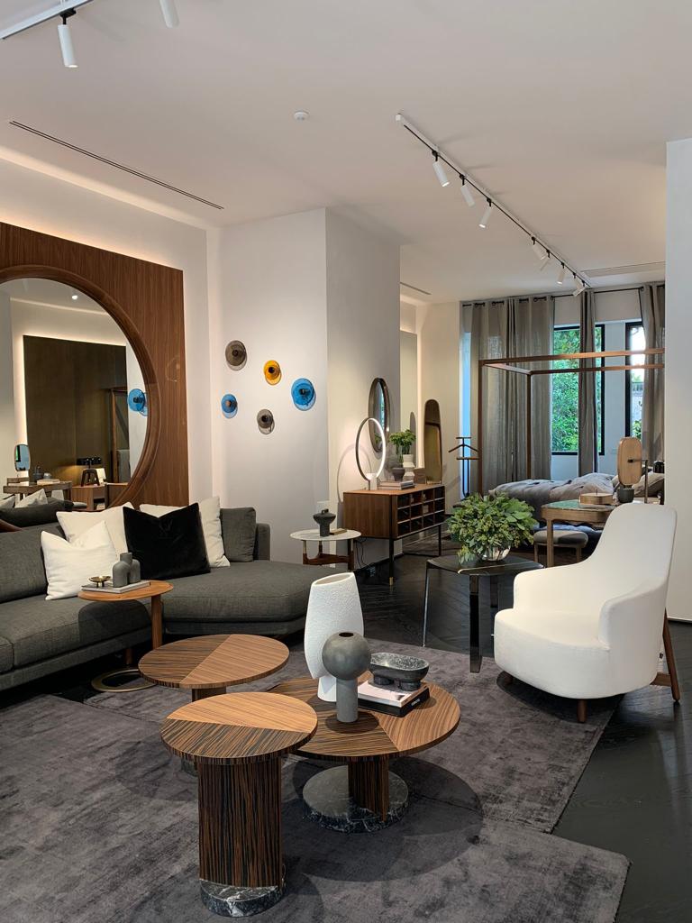 A wide-angle photo captures the main display area of a high-end furniture and design store. The space features a grey sectional sofa, several clustered round wooden coffee tables, a white accent chair, and a large circular mirror with colorful wall art, luxury showrooms.