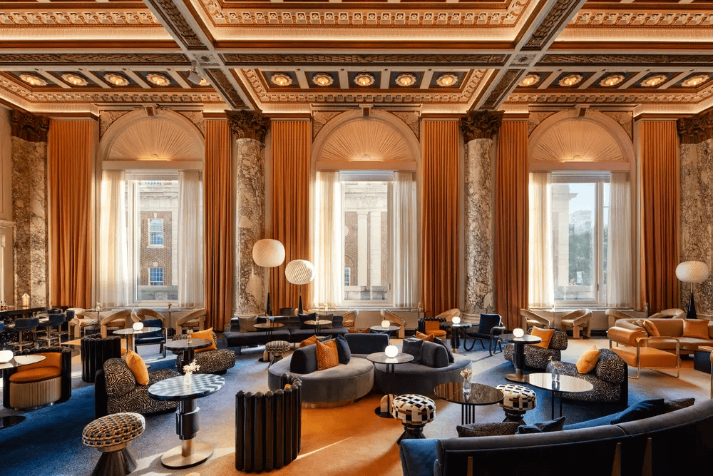 a grand, high-ceilinged hotel lounge with patterned seating arrangements, marble columns, intricate gilded detailing, and dramatic golden-orange drapery flanking tall, arched windows. ELLE Decor A-List