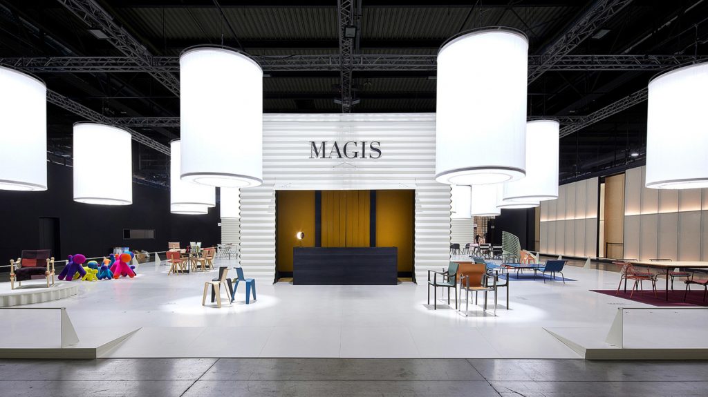 An interior view of a large, high-ceilinged trade show or gallery hall dedicated to design. Numerous large, glowing fabric cylinder lamps hang from an overhead black metal truss, illuminating diverse furniture displays, including a central reception area and surrounding seating arrangements, luxury showrooms.