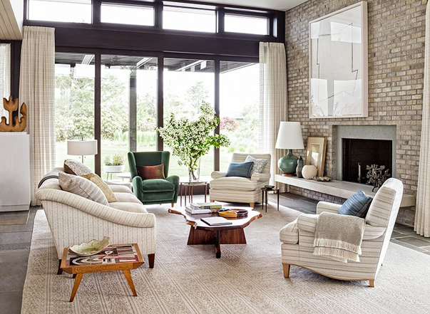 A bright, modern living room with a brick fireplace, comfortable seating, and large windows looking out to nature. ELLE Decor A-List