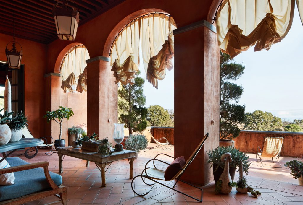 An open-air terrace with reddish walls, arched columns, fabric shades, and outdoor seating. ELLE Decor A-List