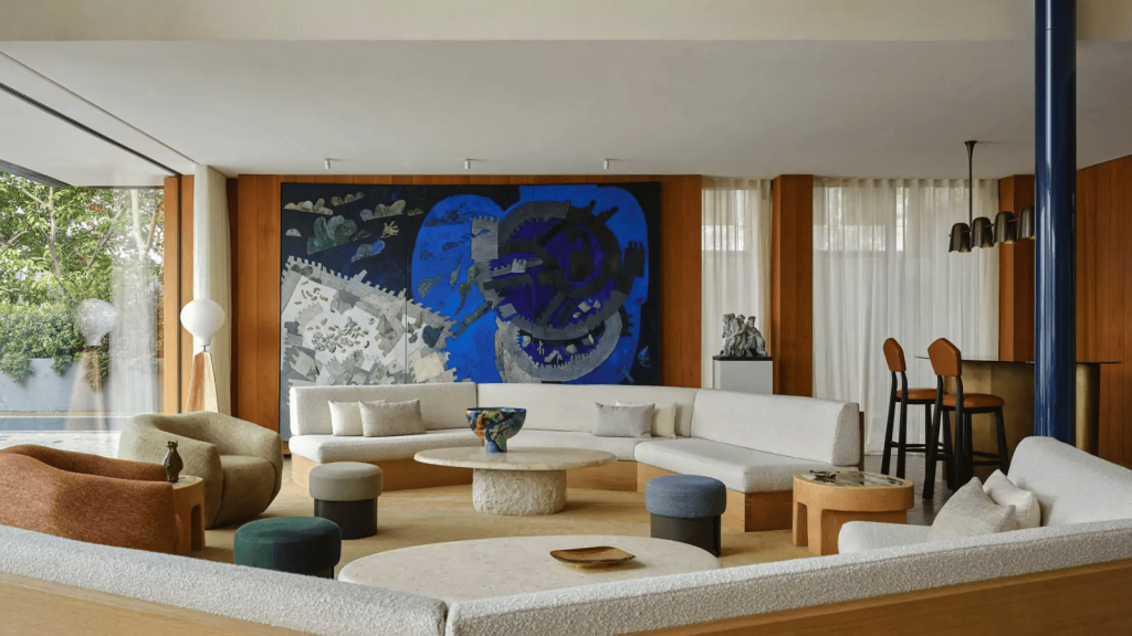 a dramatic, expansive, high-ceilinged mid-century modern space centered around a massive, curved white sectional sofa and low, round coffee tables, backed by an enormous, vibrant abstract blue mural. ELLE Decor A-List